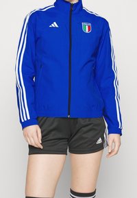 Blå idrottsjacka med vita ränder, Italien-logotyp, dragkedja fram och uppstående krage. Kombinerad med svarta shorts och randiga strumpor.