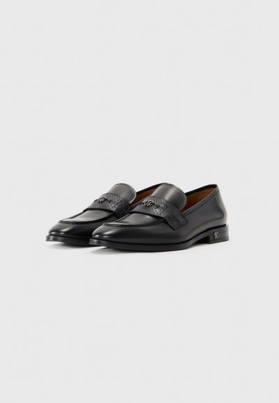 Ett par svarta läderloafers med texturerat band och små metalliska logodetaljer på loafers och häl, mot en enkel vit bakgrund.