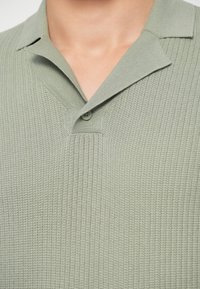 Polo shirt en tricot gaufré vert clair avec un bouton unique attaché près du col ouvert sur la partie supérieure de la poitrine d'une personne.