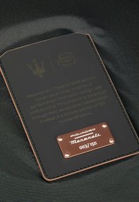 Schwarze Lederbrieftasche mit eingeprägtem Text, Kupferakzenten und einer nummerierten Plakette "003/150". Verfügt über elegante Nähte und ein minimalistisches Design.