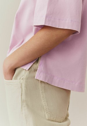 Persona valkā gaiši violeto īsroku kreklu ar roku biksēs kabatā, attēls ir apgriezts, lai parādītu vidukļa un rokas daļu.