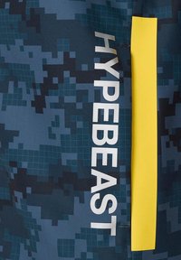 Niebieska tkanina w kamuflażu z pionowym żółtym akcentem i białym napisem "HYPEBEAST". Tekstura wydaje się gładka i syntetyczna.