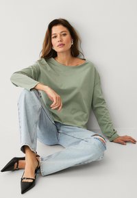 Langärmliges, weiches grünes Baumwollhemd mit weitem Ausschnitt, kombiniert mit hellblauen kaputten Jeans und schwarzen spitzen Absatzschuhen.