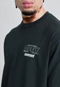 T-shirt noir en coton avec un col rond, des manches courtes et un texte imprimé blanc : "OBEY" et "movement" en polices en gras.