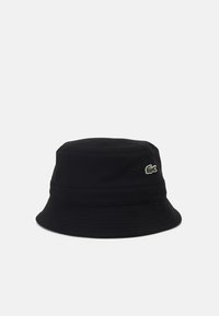 Svart bøttehatt laget av teksturert stoff, med en rund krone og vid, nedoverbøyd skjerm. Inkluderer en liten, grønn Lacoste-logo.