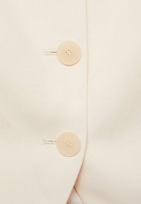 Nabijheid van beige stof met twee ronde, lichtgekleurde knopen. De textuur is glad, met een subtiele glans en zichtbare stiksels rond de knopen.