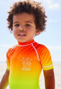 Short-sleeve, gradient rash guard in bright orange to yellow. Features white palm tree and surfboard design. Smooth, stretchy fabric.