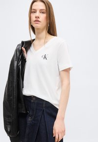 Νεαρή γυναίκα που φοράει λευκό T-shirt Calvin Klein με V-λαιμόκοψη και σκουρόχρωμη φούστα από τζιν, κρατώντας μαύρο δερμάτινο σακάκι στον ώμο της.