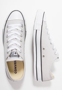 Baskets en toile grise avec embouts en caoutchouc blanc, détails de rayures noires et œillets métalliques. Les lacets sont blancs et les semelles intérieures portent le nom de la marque.