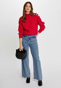 Pull en maille rouge avec un col montant, associé à un jean à jambes larges bleu clair. Sac à main noir texturé et bottes noires brillantes sont inclus.