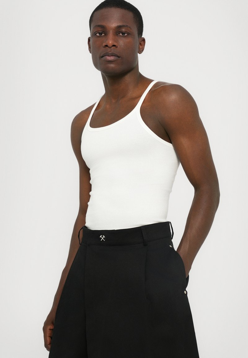 Hvid ribbet tanktop med en tætsiddende form, kombineret med højtaljede sorte bukser, der har en dekorativ detalje ved taljen.