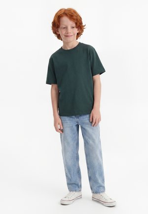 Giovane ragazzo dai capelli rossi in piedi, indossa una maglietta verde scuro, jeans azzurri e scarpe da ginnastica bianche, sorride leggermente e guarda di lato.