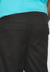 Persoon in zwarte broek en een lichtblauwe shirt, vanaf de taille naar beneden weergegeven, waarbij de achterzakken en de textuur van de stof worden benadrukt.