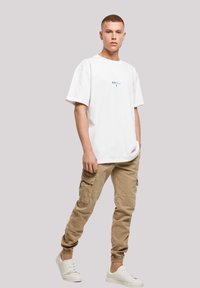 White cotton t-shirt with short sleeves and blue text, paired with light beige cargo pants featuring side pockets and elastic cuffs. White sneakers.