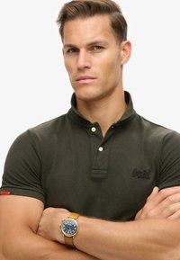 Mørkegrøn polo-shirt med klassisk krave, der har tre knapper og et logo. Modellen bærer et guldur med en brun læderrem.