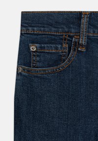Tela de mezclilla de un azul oscuro que presenta un bolsillo frontal, costuras de cobre y un botón metálico con detalle de logo. Textura suave con un diseño clásico.