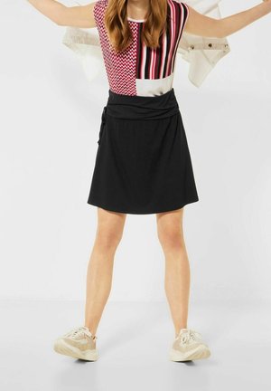 Černá zavinovací sukně s elastickým pasem, doplněná o vícbarevný top s geometrickým vzorem a béžové tenisky. Hladká textura.