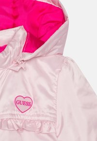 Růžová saténová bunda s kapucí, která má růžovou podšívku, volánkový lem a nášivku ve tvaru srdce s logem "GUESS".