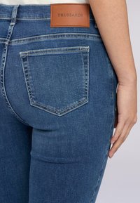 Jeans de mezclilla azul oscuro con bolsillos traseros bordados y una etiqueta de marca de piel marrón etiquetada "TRUSSARDI". La tela tiene una textura suave.