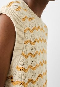 Maglione senza maniche color crema realizzato a maglia con motivi a onda testurizzati in giallo e crema alternati. Presenta un collo e delle aperture per le braccia a costine.