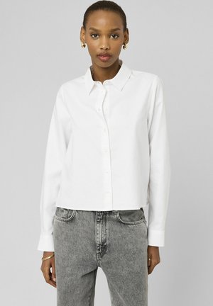 Femme aux cheveux courts portant des boucles d'oreilles créoles en or, une chemise blanche boutonnée, un jean taille haute gris, et des bracelets en or sur un fond gris.