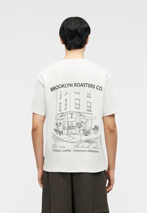 Άτομο που φοράει λευκό μπλουζάκι με μαύρη γραμμική σχεδίαση ενός καφέ σε γωνία και κείμενο "Brooklyn Roasters Co. Δυνατός καφές - Δυναμικότερες γνώμες."