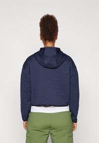 Marineblå quiltet crop hoodie med teksturert mønster, justerbar hette og ribbete nederkant. Brukt over en hvit skjorte og grønne cargobukser.