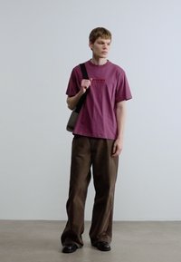 Μπορντό βαμβακερό T-shirt με μικρό λογότυπο, συνδυασμένο με χαλαρά καφέ παντελόνια κορντούρο και μαύρα παπούτσια. Διαθέτει τσάντα ώμου.