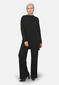 Zwarte lange mouw tuniek met een hoge nek, gecombineerd met zwarte wijde broek en hoge hakken. Het materiaal lijkt soepel en vloeiend.
