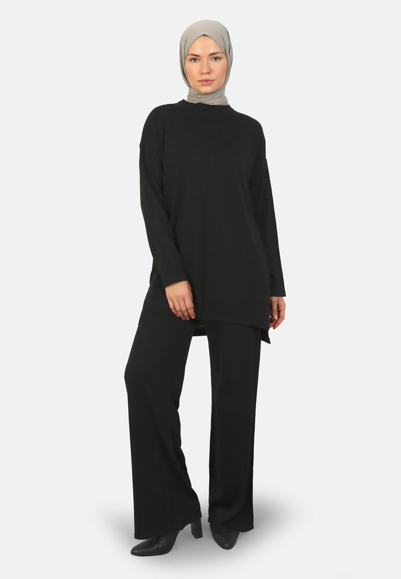 Zwarte lange mouw tuniek met een hoge nek, gecombineerd met zwarte wijde broek en hoge hakken. Het materiaal lijkt soepel en vloeiend.
