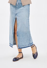 Zapatos planos azul marino con diseño de tiras trenzadas, suela marrón y correa al tobillo. Combinados con una falda de mezclilla azul clara con abertura frontal.