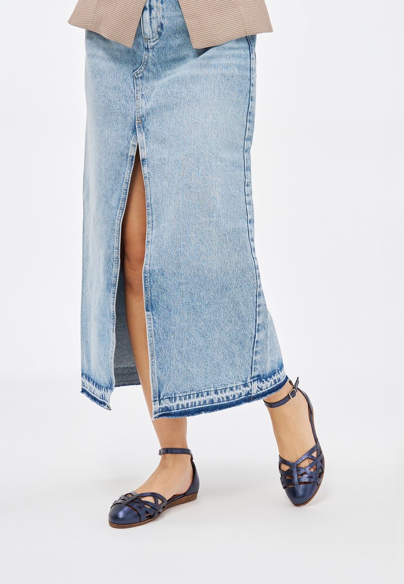 Zapatos planos azul marino con diseño de tiras trenzadas, suela marrón y correa al tobillo. Combinados con una falda de mezclilla azul clara con abertura frontal.