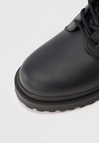 Botină din piele neagră cu vârf rotunjit, suprafață texturată, șireturi negre și o talpă robustă din cauciuc cu un model de aderență accentuat.