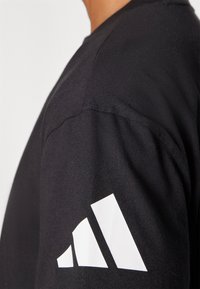 Černé bavlněné tričko s krátkým rukávem a bílým logem Adidas na levém rukávu. Hladká textura a klasický design.