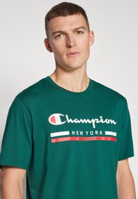 Hombre joven con el cabello corto que lleva puesta una camiseta verde de Champion New York con texto gráfico rojo y blanco en el pecho.
