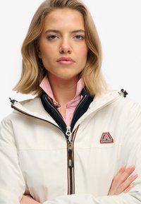 Kremowa kurtka z kapturem, z czarną warstwą wewnętrzną, pomarańczowym akcentem na suwak oraz logo góry na piersi.