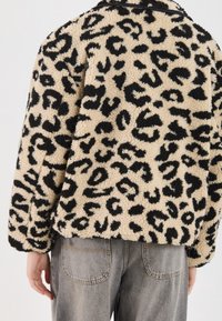 Kurtka z nadrukiem w panterkę, wykonana z pluszowego futra sztucznego w beżowej podstawie i czarnymi plamami; ma luźny krój i opuszczone ramiona. Połączona z szarymi dżinsami.