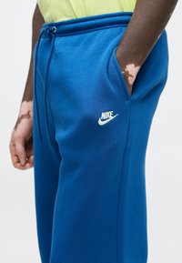 Μπλε φόρμα της Nike με ελαστική μέση και κορδόνι. Διαθέτει πλαϊνές τσέπες και μικρό λευκό λογότυπο στο αριστερό μηρό. Μαλακή υφή.