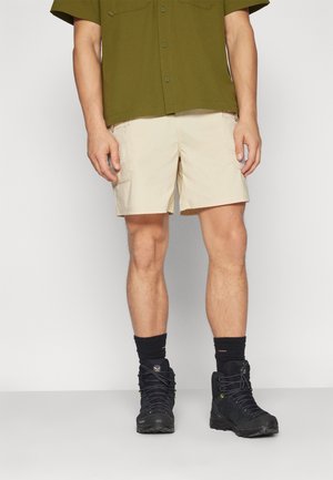 Man die beige cargoshorts draagt, zwarte wandelschoenen met zwarte sokken en een olijfgroen T-shirt met korte mouwen, staand tegen een effen achtergrond.