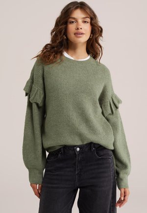 Femme aux cheveux ondulés portant un pull en maille vert olive avec des volants sur les manches et un jean noir taille haute sur un fond neutre.