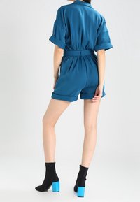 Teal kort jumpsuit med rullade ärm, elastisk midja och skräddarsydd passform. Bärs tillsammans med svarta ankelstövlar med blå klackar.