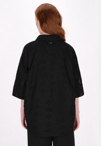 Schwarze Bluse mit Kragen, kurzen Ärmeln und lasergecuttetem Lochmuster. Weicher Stoff und lockere Passform, mit einer sichtbaren Naht im Rücken.