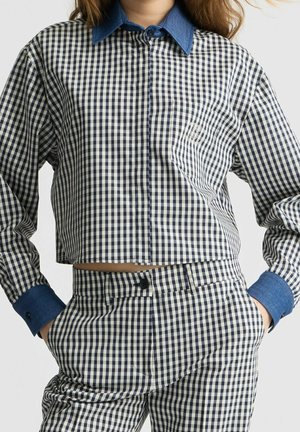 Mujer con camisa a cuadros en blanco y negro de manga larga con cuello azul y pantalones a juego con cuadros, con las manos en los bolsillos.
