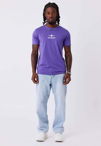 Camiseta de algodón púrpura con un logo blanco en el pecho, combinada con jeans de mezclilla azul claro y zapatillas blancas.