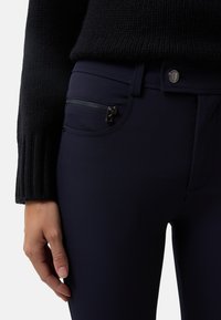 Detailní záběr na osobu, která má na sobě tmavomodré kalhoty s bočním zipovým kapsou a černý pletený svetr, ruka odpočívající u stehna.