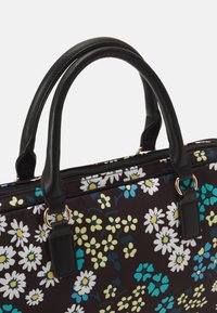 Schwarze Ledergriffe ragen von einer braunen Tasche mit floralem Muster hervor, die weiße Gänseblümchen und türkisfarbene Akzente vor einem dunklen Hintergrund zeigt.