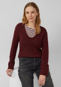 Frau mit hellbraunen Haaren, die einen kastanienbraunen gestrickten V-Ausschnitt-Pullover über einem weiß- und kastanienbraun gestreiften Shirt trägt und mit der Hand auf einer weißen Oberfläche posiert.