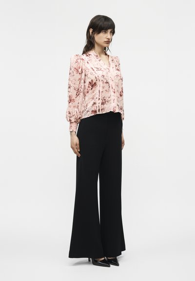 Femme portant un chemisier rose à fleurs à manches longues et un pantalon noir taille haute à jambes larges, avec des talons pointus noirs, debout devant un fond uni.