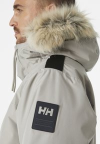 Lichtgrijze jas met een met bont gevoerde capuchon, voorzien van een zwart patch met "HH." Inclusief trekkoorden en een gestructureerde afwerking.