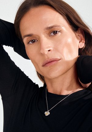 Mujer con cabello castaño que lleva un top negro y un collar con colgante en forma de corazón, posando con un brazo levantado detrás de la cabeza.